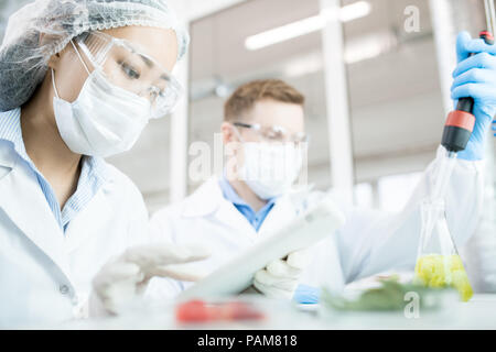 Ritratto di due scienziati moderni indossare maschere lavorando sulla ricerca alimentare studiando liquidi nel bicchiere mentre è seduto a tavola in laboratorio Foto Stock