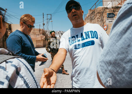 Un palestinese tour guida parla di fronte a un ingresso di insediamento a Hebron, West Bank, mentre un Forze di Difesa Israeliane si affaccia su Foto Stock