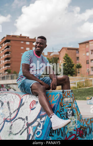 Giovane uomo in skatepark seduto sul muro, ridendo Foto Stock