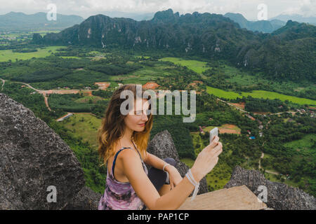 Laos, Vang Vieng, giovane donna sulla parte superiore delle rocce prendendo un selfie Foto Stock