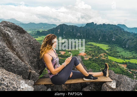 Laos, Vang Vieng, giovane donna sulla parte superiore delle rocce mediante telefono cellulare Foto Stock