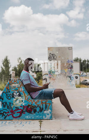 Giovane uomo in skatepark seduta sul banco, ascolto di musica Foto Stock