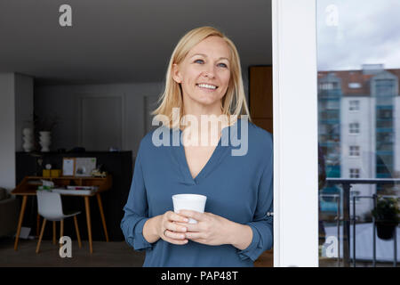 Donna sorridente holding tazza di caffè guardando fuori della porta del balcone Foto Stock