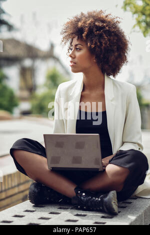 Moda giovane donna con capelli ricci seduta sul banco di lavoro con computer portatile Foto Stock