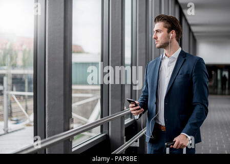 Giovane imprenditore in un passaggio con un telefono cellulare, gli auricolari e la valigia scorrevole Foto Stock