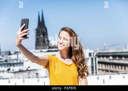 Germania, Colonia, ritratto di donna sorridente tenendo selfie con lo smartphone sulla terrazza sul tetto Foto Stock