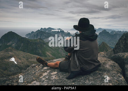 Norvegia Lofoten, Moskenesoy, giovane uomo seduto al Hermannsdalstinden, guardando sopra Kjerkefjord Foto Stock