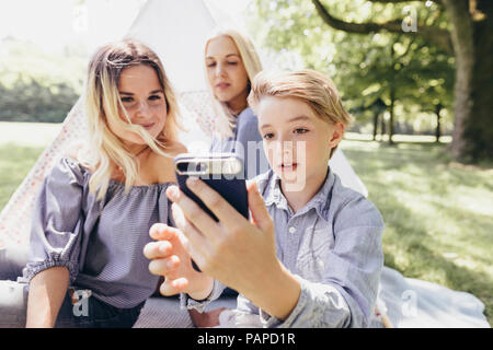 Due giovani donne e un ragazzo prendendo un selfie accanto a teepee in un parco Foto Stock