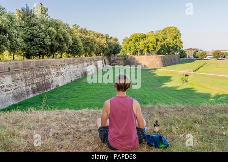 Persona con una bottiglia di birra seduti sulle antiche mura di Lucca, Toscana, Italia, in ammirazione del paesaggio circostante Foto Stock