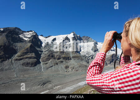 Austria, Carinzia, donna che guarda attraverso il binocolo puntando al Großglockner picco e del ghiacciaio Pasterze, vista da Kaiser-Franz-Josefs-Hoehe, Alta Tau Foto Stock