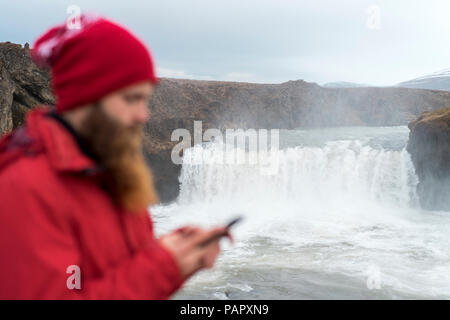 In Islanda, a nord di Islanda, giovane uomo utilizza lo smartphone, cascata in background Foto Stock