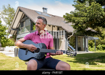 Sorridente uomo maturo seduti nel giardino della sua casa a suonare la chitarra Foto Stock