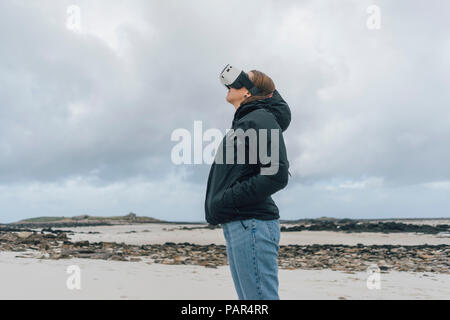 Francia, Bretagna, Landeda, giovane donna in piedi presso la costa indossando occhiali VR Foto Stock
