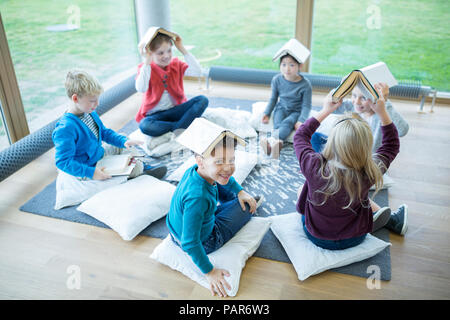 Gli alunni sorridente seduto sul pavimento nella scuola sala pausa bilanciamento libri sulle loro teste Foto Stock