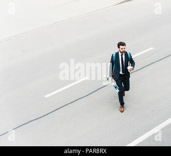 Imprenditore con caffè da asporto e skateboard a camminare sulla strada Foto Stock