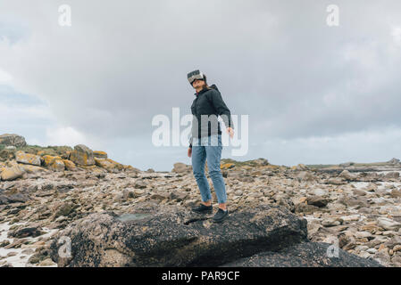 Francia, Bretagna, Landeda, giovane donna in piedi presso la costa indossando occhiali VR Foto Stock