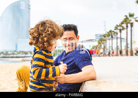 Spagna, Barcellona, giovane ragazzo giocando con sabbia, suo padre seduto accanto a lui e sorridente Foto Stock