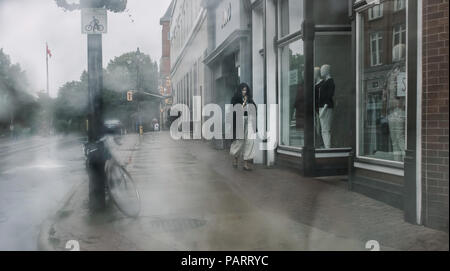 Piove a Stratford, una donna decisamente in fretta, protegge la sua testa e cammina veloce, mentre gettando uno sguardo alla vetrina di un negozio. Foto Stock