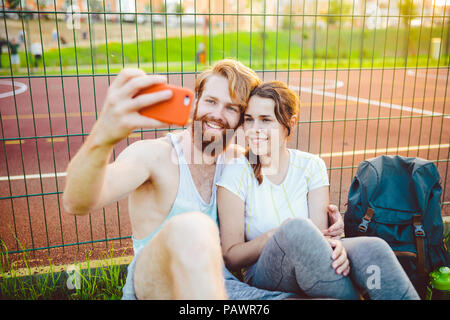 Una coppia di Le mans capelli rossi e la barba e la donna del riposo dopo la pratica di sport all'aperto sull'erba in background di Stadium. Il ragazzo tiene red smartphone su outstre Foto Stock