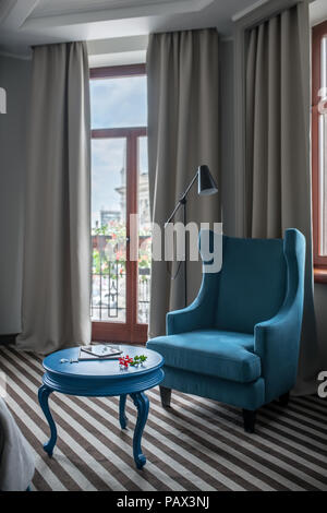 Hotel luminoso camera con pareti di colore chiaro e un pavimento striato. Vi è un giro di tavolo blu con una poltrona, finestre con tende e porte di vetro del balcone, Foto Stock