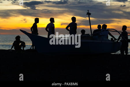 In piedi e seduta gruppo di pescatori con barca da pesca silhouette isola sulla spiaggia al tramonto - Panay, Filippine Foto Stock