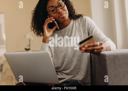 Donna seduta su un divano a casa parlando al telefono cellulare in possesso di una carta di credito con un computer portatile sul suo grembo. Donna fare il pagamento on-line sul suo laptop comput Foto Stock