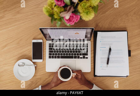 Vista dall'alto di un imprenditore donna seduta con il computer portatile e il telefono cellulare sulla tabella tenendo una tazza di caffè in mano. Imprenditrice di lavoro a casa wi Foto Stock