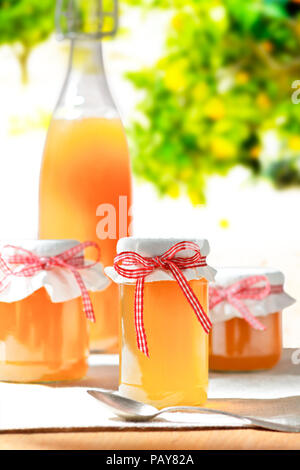 In casa succo di frutta in una bottiglia di vetro e jelly in vasi con coperchio di lino e un nostalgico ribbon bow nel sole splendente, alberi da frutto in background Foto Stock