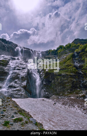 Cascata di montagna sotto un cielo nuvoloso, illuminata dai raggi del sole con ghiacciaio su un primo piano Foto Stock
