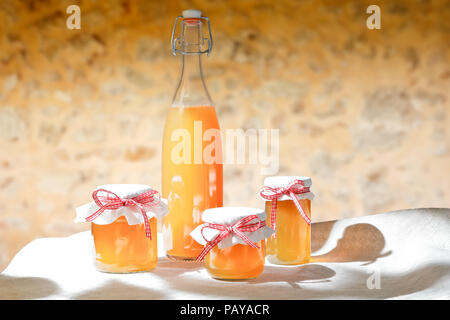 In casa succo di frutta in una bottiglia di vetro e jelly in vasi con coperchio di lino e un nostalgico ribbon bow nel sole splendente nella parte anteriore di un rustico in pietra. Foto Stock