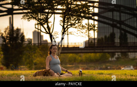 Bella incinta donna giovane con little dog sitter su mat, godendo di yoga, rilassante, sentirsi vivi, respirando aria fresca, calma e sognare, nel verde parco di estate. Foto Stock