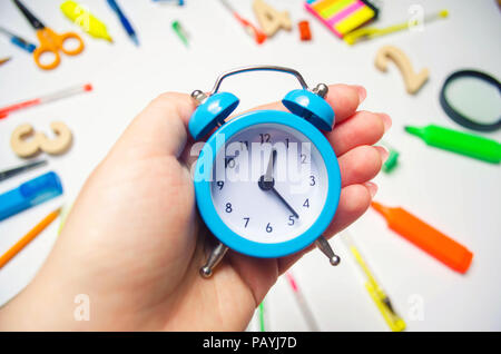 Si torna a scuola. blu sveglia sul banco di scuola nelle mani di uno studente. cancelleria. accessori. Sfondo bianco. adesivi, penne colorate, penc Foto Stock