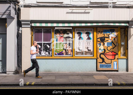 Uomo che cammina in Old Compton Street nel quartiere di Soho di fronte ad una parete ricoperta di cartelloni murali e Foto Stock