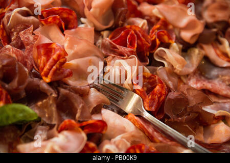 Close up di diversi tipi di carne italiana (ham) su una piastra con una forcella e una foglia di basilico Foto Stock