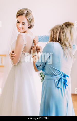 La vista laterale del bridesmaids aiutando la sposa bsmiling con l'abito da sposa. Foto Stock