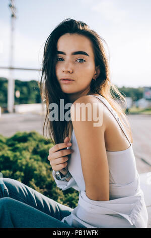 Piuttosto giovane donna che indossa top bianco e jeans seduti all'aperto al giorno d'estate e di sole. Ragazza con lunghi capelli castani e gli occhi blu che pongono guardando la fotocamera. Foto Stock