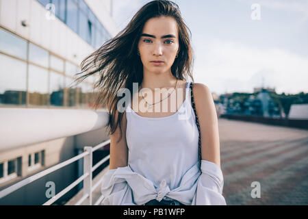 Giovane e bella donna che indossa top bianco che pongono a city street al giorno d'estate. Ragazza con svolazzanti capelli lunghi marrone in piedi all'esterno guardando la fotocamera. Foto Stock