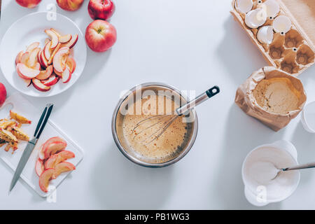 Ingredienti e strumenti per cucinare la torta di mele, vista dall'alto. Recipiente metallico riempito con uova di panna montata, frusta e tagliare la frutta, gusci d'uovo, farina e zucchero, laici piatto co Foto Stock