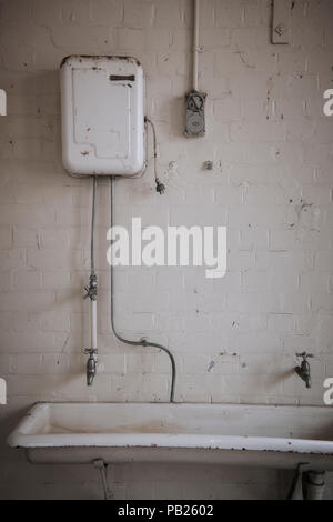 Vecchio abbandonato acqua calda servizio su un grande porcellana bianca lavabo in un mattone bianco camera. Foto Stock