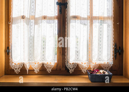 Una finestra in legno con un bianco cortina ricamata e un piccolo cestino di fiori secchi sul davanzale Foto Stock