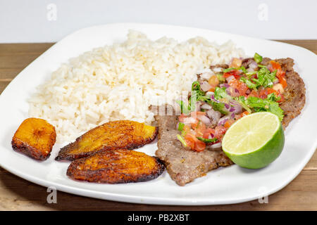 Una bistecca coperto di Pico de Gallo circondato da platani e riso bianco su una piastra bianca. Cibo cubano. Foto Stock