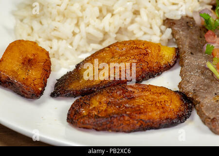 Una bistecca coperto di Pico de Gallo circondato da platani e riso bianco su una piastra bianca. Cibo cubano. Foto Stock