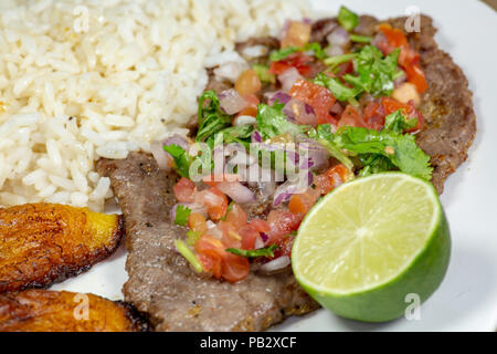 Una bistecca coperto di Pico de Gallo circondato da platani e riso bianco su una piastra bianca. Cibo cubano. Foto Stock