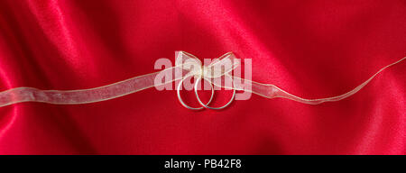Una coppia di golden fedi legate con un nastro d'oro su raso rosso sfondo, banner Foto Stock