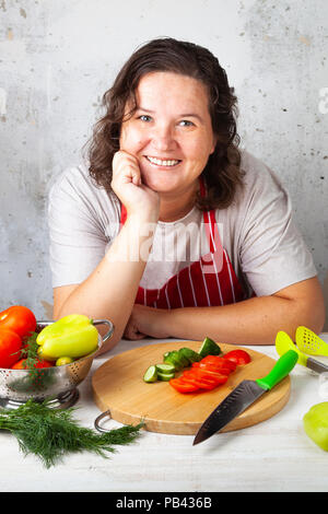 La donna in un grembiule rosso si siede a tavola in cucina e sorrisi. Di mezza età casalinga che prepara la cena. Foto Stock