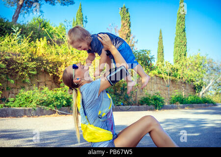 Famiglia praticare sport insieme, bella madre sportive facendo esercizi nel parco sul tappetino con il suo grazioso piccolo figlio, sollevando la sua preziosa baby Foto Stock