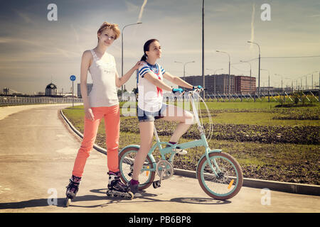 Due giovani amici di sesso femminile uno su una bicicletta, uno su un scates del rullo. Estate Sport e tempo libero tema. Dai toni caldi immagine Foto Stock