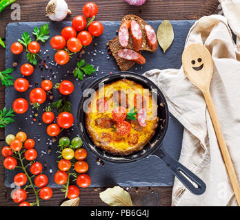 Fritti frittata di uova di pollo con rossi pomodori ciliegini e salsiccia affumicata in un tondo nero padella su un tavolo nel mezzo di verdure, top v Foto Stock