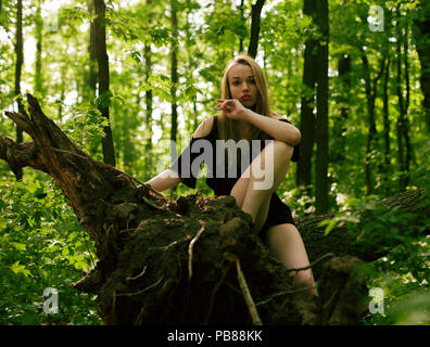 Ragazza in un bosco di latifoglie siede su un albero caduto in estate su una luminosa giornata di sole Foto Stock