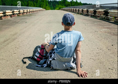 Hitchhiker. Adolescente viaggiatore con uno zaino seduto sul pavimento su una autostrada deserta. Inquadratura orizzontale. Extreme concetto di turismo Foto Stock